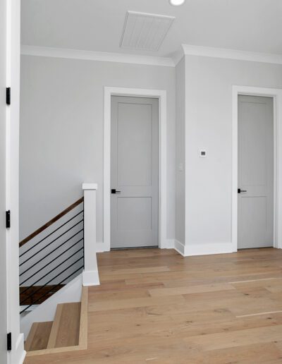 A modern hallway with two closed white doors, light hardwood floors, and a glimpse of a staircase with metal railings.